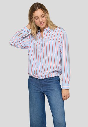 Woman wearing light blue blouse with red and white vertical stripes and blue jeans, posing with one hand touching her hair.