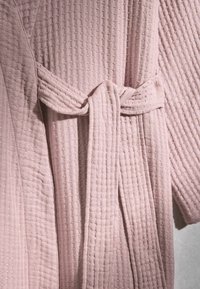 Peignoir en tissu texturé rose clair avec une ceinture nouée à la taille, montrant des motifs carrés matelassés sur le matériau.