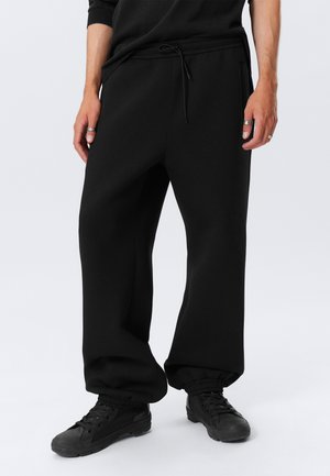 Homme portant un pantalon de survêtement noir ample avec des poignets élastiques, des baskets noires et un haut noir, debout les mains détendues le long du corps.