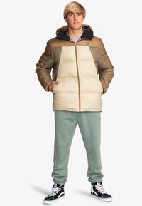 Pufferjack van beige en dof bruin met een capuchon, met een ritsluiting en gequilt textuur. Gecombineerd met groene sweatpants en zwarte sneakers.