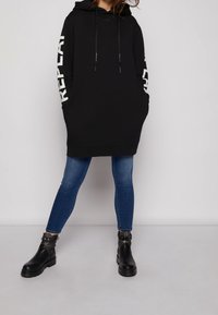 Person som bär en svart oversize hoodie med "REPLAY" text på ärmarna, blå skinny jeans och svarta ankelstövlar, står mot en vit bakgrund.