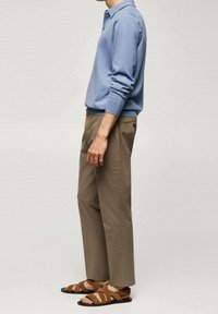 Uomo che indossa una camicia a maniche lunghe blu, pantaloni cropped marroni e sandali di pelle marroni, in piedi su uno sfondo semplice.