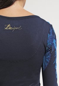 Granatowa bluzka z długim rękawem z niebieskim kwiatowym wzorem na rękawach, okrągłym dekoltem i złotym haftowanym logo "Desigual" z tyłu.