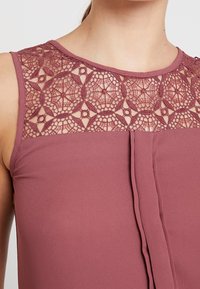 Mauve blůza s krajkovým yoke s složitými geometrickými vzory, jemný materiál, bezrukávový design a plisovaný přední detail.