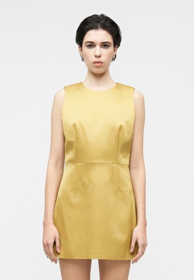 Femme aux cheveux courts et foncés portant une robe mini jaune en satin sans manches, debout devant un fond blanc uni, regardant vers l'avant.