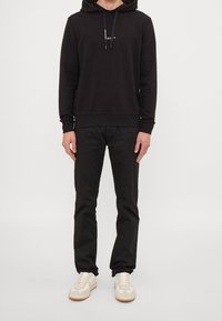 Felpa nera con cappuccio, lacci e logo frontale, abbinata a pantaloni neri. Il modello indossa sneakers chiare con accenti beige. Outfit semplice e casual.