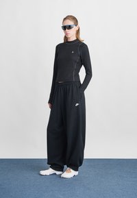 Naine seisab sinisel vaibal, seljas must pikka varrukaga topp, üle mõõtu mustad Nike dressipüksid, valged slip-on kingad ja sinised ümmargused prillid.