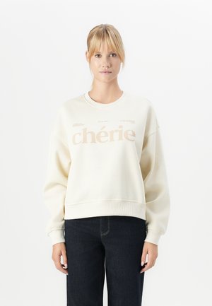 Jeune femme aux cheveux blonds portant un sweat-shirt crème avec le texte "chérie" et un jean foncé, se tenant devant un fond blanc uni.