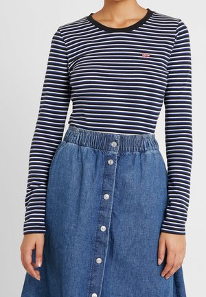 Femme portant un haut à manches longues rayé noir, blanc et bleu, associé à une jupe en denim bleue taille haute avec boutons sur le devant.