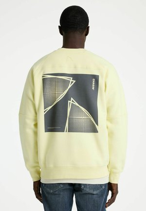 Person von hinten, trägt ein hellgelbes Sweatshirt mit schwarz-weißem geometrischem Muster und dem Schriftzug "CHASIN'" auf dem Rücken, kombiniert mit blauen Jeans.