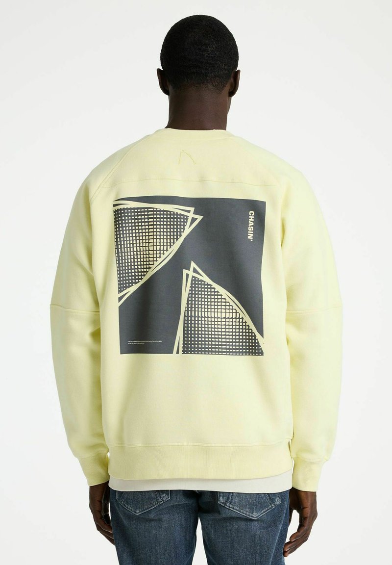 Person von hinten, trägt ein hellgelbes Sweatshirt mit schwarz-weißem geometrischem Muster und dem Schriftzug "CHASIN'" auf dem Rücken, kombiniert mit blauen Jeans.