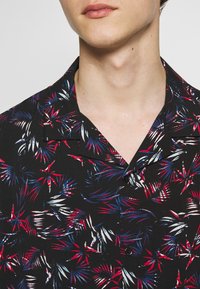 Schwarzes Kurzarmhemd mit tropischem Blattmuster in Rot, Weiß und Blau. Verfügt über einen Camp-Kragen und eine Knopfleiste vorne.