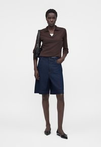 Femme portant un haut marron à manches longues, un short en jean foncé jusqu'aux genoux, des ballerines noires et un sac à épaule noir, debout avec une main dans la poche.