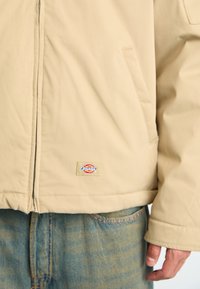 Beige zip-up jacket with side pockets worn over blue denim jeans, showing a small Dickies logo patch near the jacket hem.