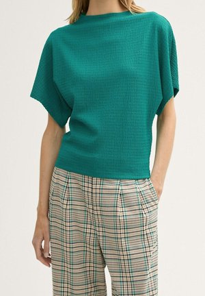 Frau trägt ein grünes, strukturiertes Kurzarmshirt kombiniert mit hoch taillierten beigen karierten Hosen, die Hände in den Taschen, vor einem einfarbigen Hintergrund.