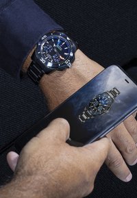 Melns nerūsējošā tērauda pulkstenis ar zilu ciparnīcu un sudraba akcentiem; viedtālrunis parāda līdzīgu pulksteņa dizainu. Rokas pulkstenim ir hronogrāfa detaļas.