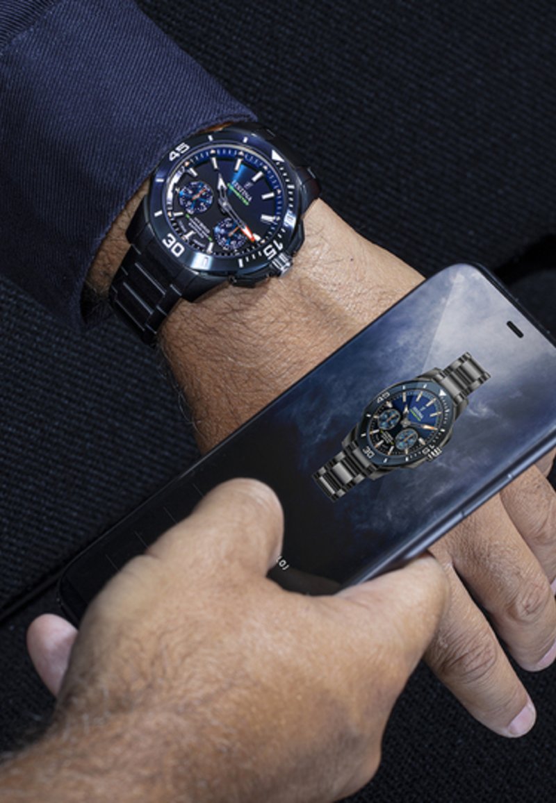 Melns nerūsējošā tērauda pulkstenis ar zilu ciparnīcu un sudraba akcentiem; viedtālrunis parāda līdzīgu pulksteņa dizainu. Rokas pulkstenim ir hronogrāfa detaļas.