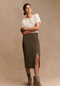 Blouse blanche froissée avec fermeture à boutons, associée à une jupe olive longueur genou avec une fente sur le côté et des bottes en cuir serpent marron texturé.