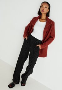 Red textured coat, white ribbed top, black striped wide-leg pants, black and burgundy block-heeled shoes. Sanitary setting with neutral background.