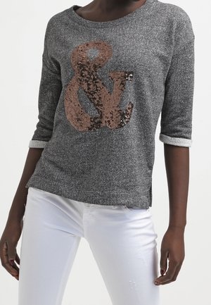 Persoon die een grijs shirt met lange mouwen en opgerolde manchetten draagt, met een groot met pailletten versierd ampersand-teken, gecombineerd met witte broek.