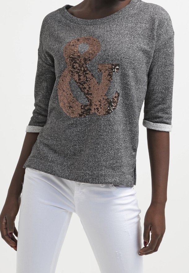 Personne portant une chemise grise à manches longues avec des poignets retroussés et un grand symbole esperluette en sequins, associée à un pantalon blanc.