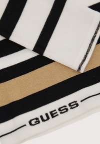 Écharpe rayée avec des couleurs alternées blanc, noir et beige. Le bord fini présente le logo "GUESS" en noir sur un fond blanc. Texture douce.