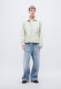 Απαλό πράσινο βαμβακερό μπουφάν με κουμπιά στο μπροστινό μέρος, δύο τσέπες και άνετη εφαρμογή, συνδυασμένο με φαρδιά ανοιχτό μπλε τζιν τζιν. Μαύρα παπούτσια.