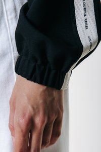 Manga de tela negra con puño fruncido, con una franja blanca en contraste impresa con "COLOURFUL REBEL". Textura suave, diseño atlético.