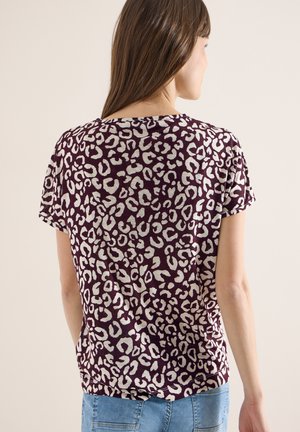 Femme aux cheveux bruns raides, portant un t-shirt à manches courtes violet à imprimé léopard et un jean bleu clair, vue de dos sur un fond uni.