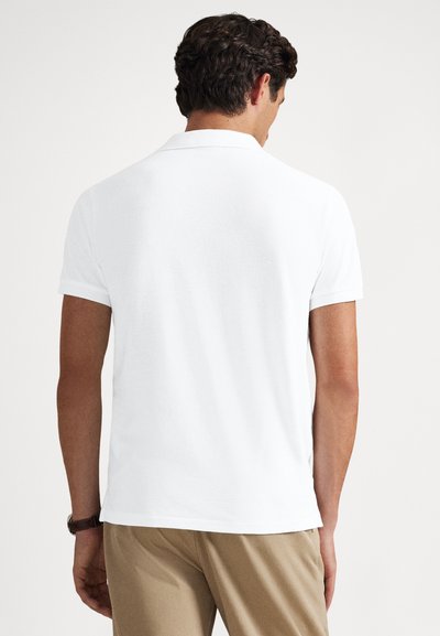 Polo branca de manga curta, design simples, textura suave e corte ajustado. Usada com calças cáqui, vista de costas.