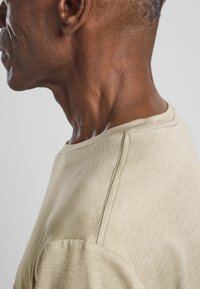 Beige Kurzarmhemd mit Rundhalsausschnitt. Weicher, strukturierter Stoff mit dezenten Stichdetails an der Schulter und am Dekolleté.