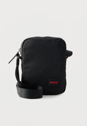 Zwarte rechthoekige crossbodytas met ritssluiting, verstelbare riem, subtiel logopatroon en rode "HUGO"-label op het voorvak.