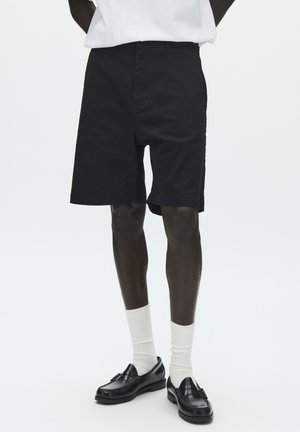 Čierne bavlnené šortky s klasickým strihom, majú rovný predný diel, bočné vrecká a dĺžku do polovice stehna, v kombinácii s bielymi ponožkami a čiernymi topánkami.