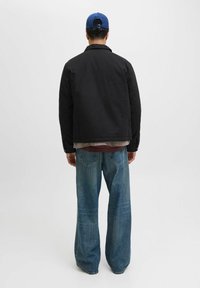 Giacca nera con una camicia sotto e un cappellino blu, abbinata a jeans larghi di un blu chiaro, vista da dietro su uno sfondo bianco.