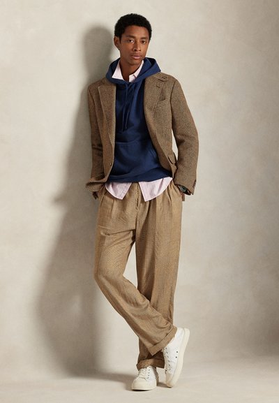 Tânăr sprijinit de perete, purtând costum maro în carouri, hanorac bleumarin, cămașă roz și adidași albi, mâinile în buzunare.