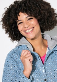 Veste en denim de couleur bleue, avec capuche grise, recouverte de sequins argentés, fermeture à bouton, et coupe ajustée.