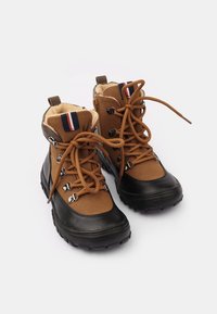 Botas marrones con suelas de goma negras, con forro de piel sintética, cordones marrones y ojales plateados. Detalle a rayas azul y rojo en el puño.