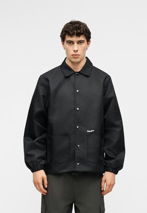 Bărbat tânăr în picioare, purtând o jachetă neagră cu nasturi, cu manșete elastice și două buzunare frontale, asortată cu pantaloni cargo închiși la culoare.