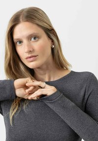 Graues Langarmoberteil mit dezentem Schimmer und weitem Ausschnitt. Das Model trägt einen silbernen Ring und runde Ohrringe, die Hände sind verschränkt.