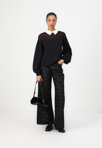 Schwarzer Pullover mit weißem Kragen, kombiniert mit weit geschnittenen schwarzen Hosen mit Muster. Trägt eine kleine schwarze Handtasche und glänzende schwarze Schuhe.