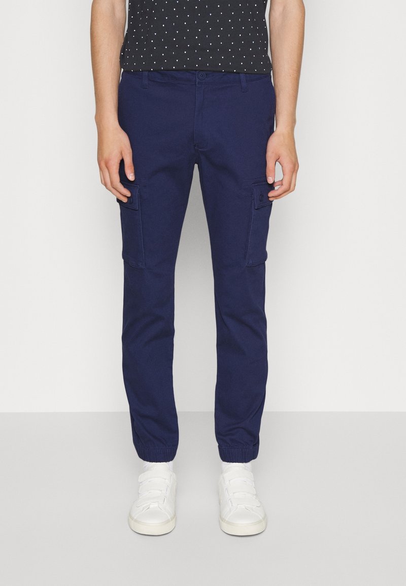 Personne portant un pantalon de jogging cargo bleu marine avec des poignets élastiques, associé à des baskets blanches à velcro et un haut noir à pois blancs.