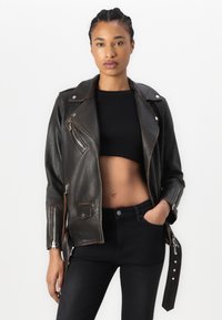 Giacca da biker in pelle nera con cerniere argentate, colletto a scatto e dettaglio con fibbia laterale. Indossata sopra un crop top nero e jeans a vita alta.