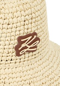 Geweven strooien emmerhoed in lichtbeige met een bruin leren logo-patch met reliëf cursieve initialen in crème. Getextureerd ontwerp met brede rand.