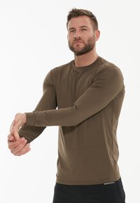 Brun langærmet sports T-shirt med rund hals, glat tekstur, tætsiddende design og sort "ENDURANCE" logo ved kanten. Model strækker armen.