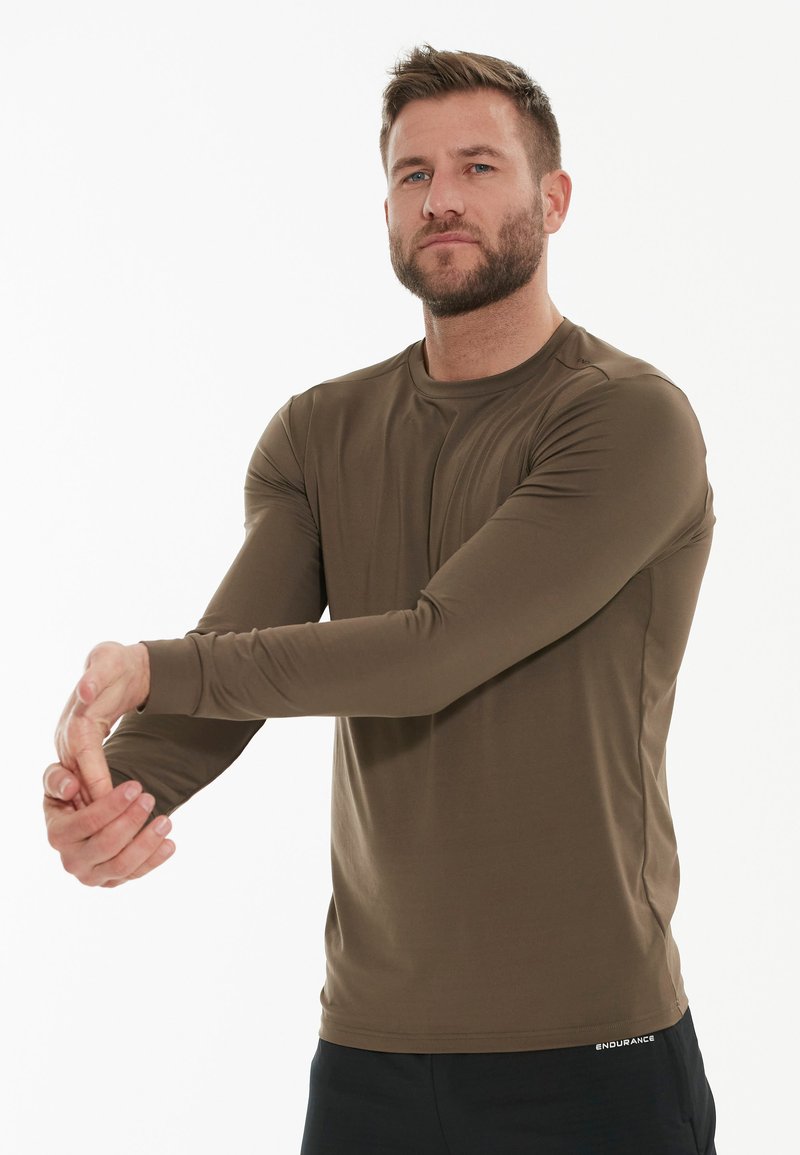 Brun langærmet sports T-shirt med rund hals, glat tekstur, tætsiddende design og sort "ENDURANCE" logo ved kanten. Model strækker armen.