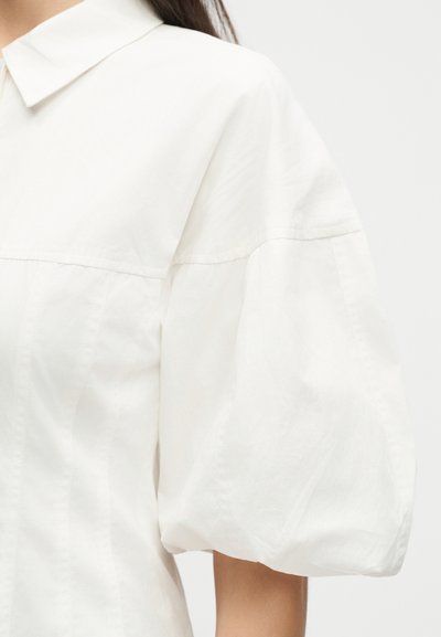 Γυναίκα που φοράει λευκό πουκάμισο με φαρδύ, φουσκωτό κοντό μανίκι και κλασικό γιακά μπροστά σε απλό φόντο.