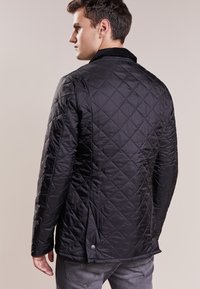 Casaco preto acolchoado com colarinho de brim, fechos de botão na parte inferior e costura diagonal texturizada. Possui mangas longas e um acabamento suave.