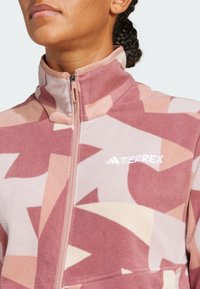 Chaqueta de forro polar con cuello alto, que presenta un patrón geométrico en rosa, beige y burdeos. Cierre con cremallera y logotipo en el pecho.