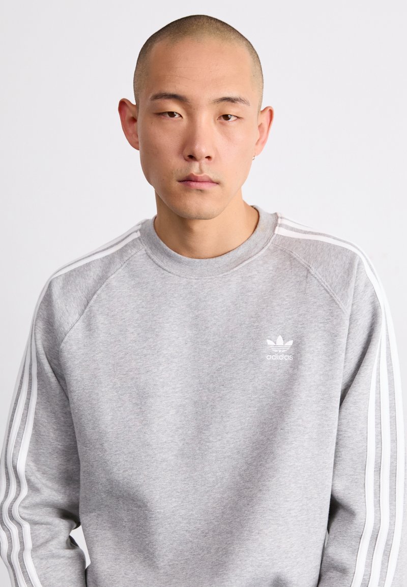 Sudadera gris hecha de material suave, con rayas blancas en las mangas y un pequeño logo blanco en el pecho. Diseño clásico de cuello redondo.