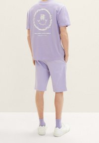 Camiseta de algodón lila y pantalones cortos a juego. La camiseta presenta un gráfico y texto en la parte posterior. Zapatillas blancas y calcetas lila completan el look.